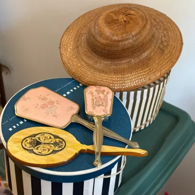 Sale Photo Thumbnail #39: Vintage vanity vanity set featuring embroidered pink floral hand mirrors and a celluloid hand mirror, displayed on striped Strawbridge & Clothier hat boxes topped with a woven straw hat.