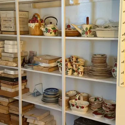 Sale Photo Thumbnail #23: White shelving filled with various kitchenware, including a fruit-themed ceramic dish set, a decorative teapot, stacked plates, and boxed items.