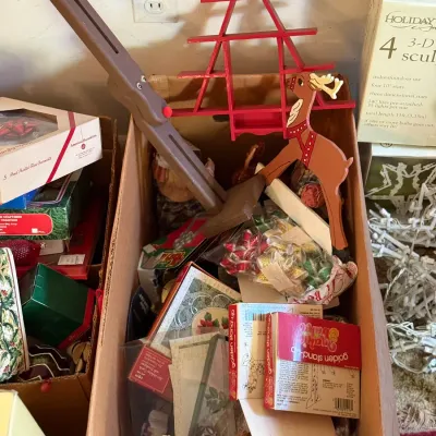 Sale Photo Thumbnail #55: Cardboard box filled with assorted vintage Christmas decor, including a red wooden tree-shaped shelf, a wooden reindeer cutout, boxed holiday cards, and ornaments.