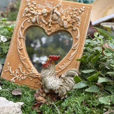 Sale Photo Thumbnail #197: Vintage resin heart-shaped mirror frame with ornate cherub details, displayed alongside a painted rooster figurine in an outdoor garden setting.