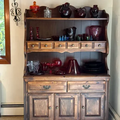 Sale Photo Thumbnail #122: Vintage dark stained pine hutch with tiered upper shelves, small apothecary drawers, and lower cabinet storage, displayed with various ruby red, amber, and clear glass serving pieces.