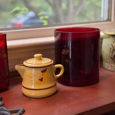 Sale Photo Thumbnail #188: Vintage yellow ceramic rooster creamer pitcher with lid, displayed on a wooden shelf alongside a deep ruby red glass cylinder vase.