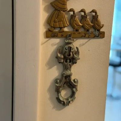 Sale Photo Thumbnail #167: Vintage brass Sunbonnet Sue and ducks key hook rack mounted on a wall with a weathered antique metal door knocker hanging below it.