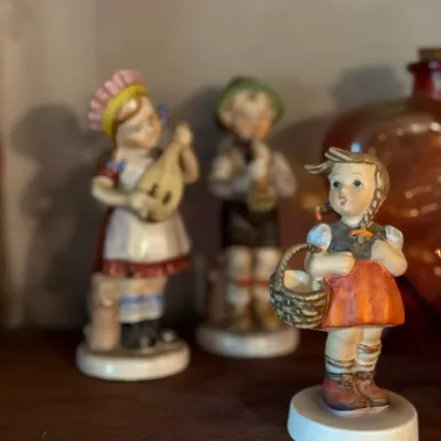 Sale Photo Thumbnail #91: Vintage Goebel Hummel porcelain figurine depicting a young girl with a basket, displayed alongside two additional Hummel figurines on a wooden shelf.