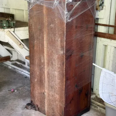 Sale Photo Thumbnail #259: Tall antique wooden cabinet wrapped in protective plastic, featuring carved feet and a warm reddish-brown finish.