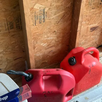 Sale Photo Thumbnail #215: Two red plastic gasoline cans with black caps sitting near a wooden wall, showing light wear and handwriting on the plastic.