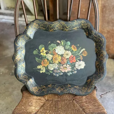 Sale Photo Thumbnail #264: Vintage black hand-painted metal tray with a scalloped edge, floral bouquet center, and gilt scrollwork border, displayed on a wooden woven-seat chair.