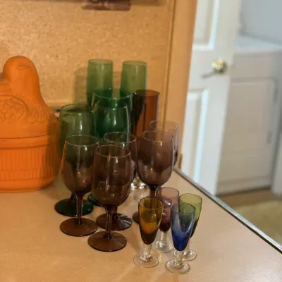 Sale Photo Thumbnail #146: Collection of mid-century colored glassware including green and smoky brown wine glasses, tall flutes, and small multi-colored cordial glasses, displayed next to a terracotta duck baker.