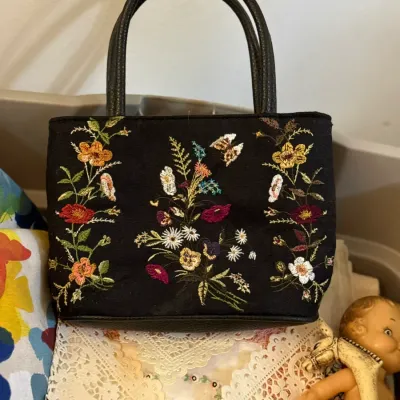 Sale Photo Thumbnail #33: Black fabric handbag featuring colorful embroidered floral motifs and dual black handles, resting on lace doilies.