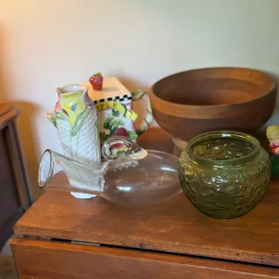 Sale Photo Thumbnail #78: Assortment of decorative tabletop items, including an olive green textured glass bowl, a hand-blown clear glass watering vessel, a floral ceramic vase, and a turned wooden bowl.
