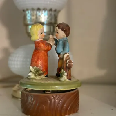 Sale Photo Thumbnail #153: Vintage ceramic music box featuring figurines of a young boy and girl standing on a faux wood-grain base.