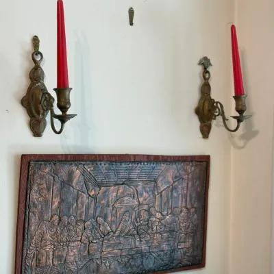 Sale Photo Thumbnail #149: Framed copper relief plaque depicting The Last Supper, displayed below a pair of vintage brass wall sconces holding red taper candles.