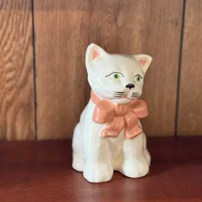 Sale Photo Thumbnail #181: Vintage white ceramic cat figurine with green eyes and a pink bow, sitting on a wooden shelf.