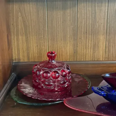 Sale Photo Thumbnail #110: Vintage ruby red glass covered butter dish in a Moon and Stars pattern, resting on a wooden cabinet shelf alongside other glassware.