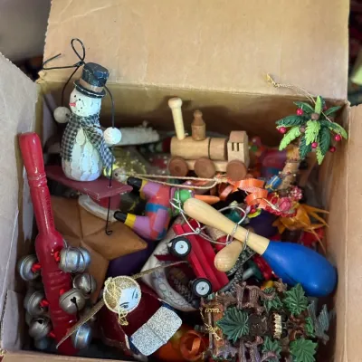 Sale Photo Thumbnail #54: Cardboard box filled with assorted vintage Christmas ornaments, including a wooden snowman on a sled, a miniature train, hand bells, and a palm tree ornament.