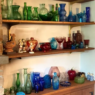 Sale Photo Thumbnail #92: Collection of vintage colored glass glassware, bottles, and ceramic figurines displayed on mid-century modern wooden shelving.