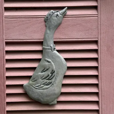 Sale Photo Thumbnail #196: Vintage cast metal goose wall plaque depicting a goose wearing a hat and ribbon collar, mounted against a pink wooden shutter.