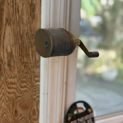 Sale Photo Thumbnail #192: Vintage hand-crank pencil sharpener mounted to a window frame, showing age-appropriate patina and wear.