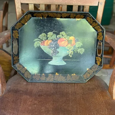 Sale Photo Thumbnail #266: Vintage octagonal black metal serving tray with hand-painted fruit still life design and gilded grape leaf border, resting on a wooden chair.