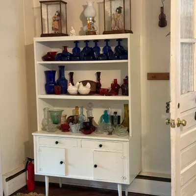 Sale Photo Thumbnail #150: White painted mid-century hutch filled with colorful vintage glassware, including cobalt blue, ruby red, and clear glass pieces, along with framed Japanese dolls on top.