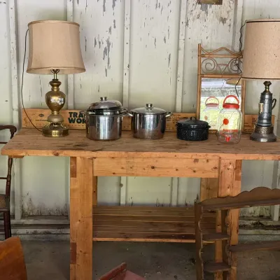 Sale Photo Thumbnail #277: Rustic wooden workbench displays vintage table lamps, stainless steel stock pots, a speckled roaster, and decorative items alongside surrounding chairs and shelving.