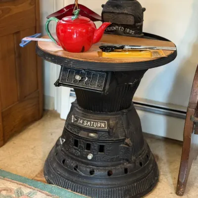 Sale Photo Thumbnail #136: Antique 14 Saturn cast iron potbelly stove repurposed as a side table with a wooden top, displayed with a red teapot and decorative items.