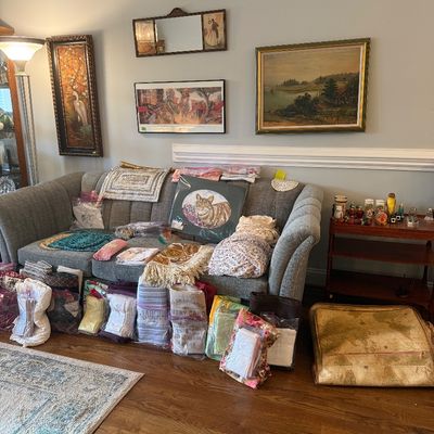 Sale Photo Thumbnail #55: Gray upholstered sofa laden with assorted textiles, linens, and framed artwork, accompanied by framed wall art and a small wooden side table.