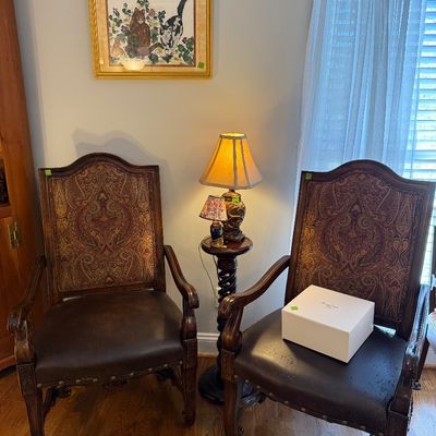 Sale Photo Thumbnail #23: Pair of Traditional upholstered armchairs with paisley backs and dark leather seats, flanked by a small accent table holding lamps, and a framed cat artwork above.