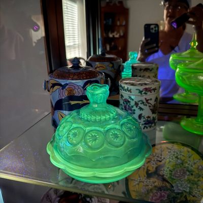 Sale Photo Thumbnail #6: Vintage green uranium glass covered butter dish in Moon and Star pattern glowing under UV light, displayed alongside decorative ceramics.