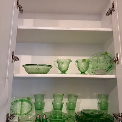 Sale Photo Thumbnail #9: Collection of green Depression glassware displayed inside a white cabinet, including tumblers, plates, bowls, a cream and sugar set, and salt and pepper shakers.