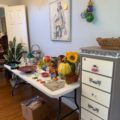 Sale Photo Thumbnail #25: A folding table displayed with various colorful home decor, ceramics, and glassware next to a painted tallboy dresser and framed wall art.