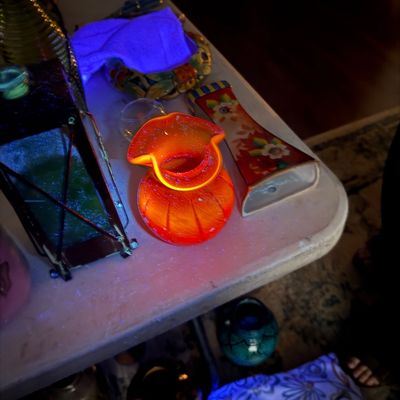Sale Photo Thumbnail #4: Collection of decorative glassware and ceramics displayed under UV blacklight, including a glowing orange-red cadmium glass vase.