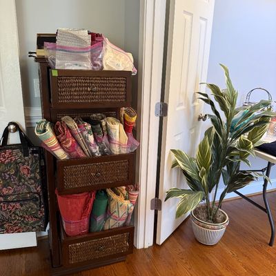Sale Photo Thumbnail #26: Tall dark wood storage tower with woven rattan drawers filled with folded linens, displayed alongside a potted artificial plant.