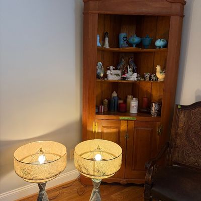 Sale Photo Thumbnail #24: Vintage wood corner hutch with swan neck pediment, displayed alongside a pair of mid-century ceramic table lamps and a carved armchair.