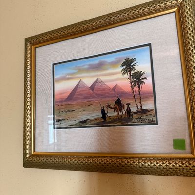 Sale Photo Thumbnail #70: Framed Egyptian desert scene painting depicting figures and a camel near the Pyramids of Giza, set in an ornate gold frame with a fabric mat.