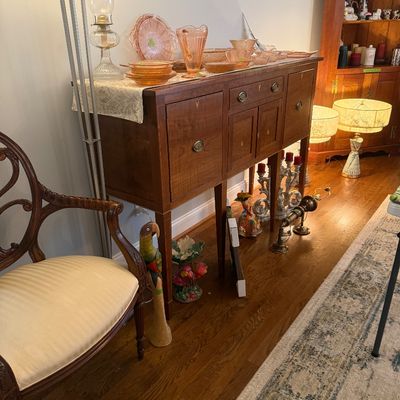 Sale Photo Thumbnail #54: Traditional wooden sideboard with inlaid details, displaying a collection of pink Depression glass tableware alongside an ornate wooden armchair.