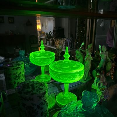 Sale Photo Thumbnail #5: Pair of vintage etched uranium glass pedestal candy dishes glowing bright green under UV light, displayed among other glassware.