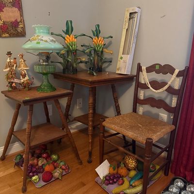 Sale Photo Thumbnail #65: Corner display featuring wooden side tables, a green glass oil-style lamp, a rush-seat ladderback chair, decorative beaded fruit, and metal banana plant sculptures.