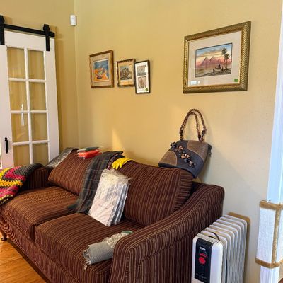 Sale Photo Thumbnail #38: Brown pinstriped upholstered sofa laden with various household linens, framed artwork on the yellow wall, and a portable oil-filled space heater on the floor.