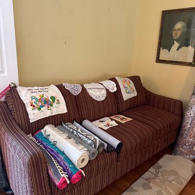 Sale Photo Thumbnail #33: Brown pinstriped upholstered sofa displayed with rolled area rugs, embroidered linens, and lace doilies, alongside a framed George Washington portrait print.