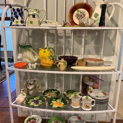 Sale Photo Thumbnail #10: White metal baker's rack display filled with various ceramic pitchers, majolica-style fruit plates, novelty teapots, and serving ware.
