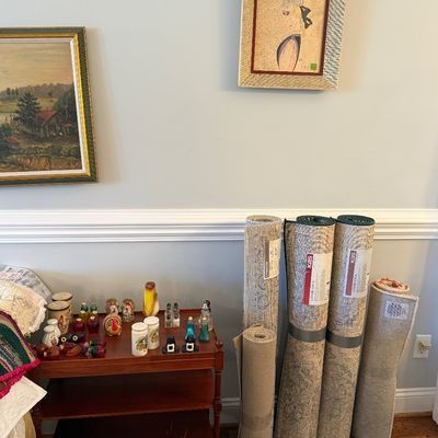 Sale Photo Thumbnail #17: Collection of rolled area rugs leaning against a wall next to a wooden cart displaying novel salt and pepper shakers, below framed wall art.