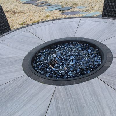 Sale Photo Thumbnail #51: Modern round outdoor patio fire pit table featuring a gray stone-tile top and blue glass fire beads, flanked by black wicker chairs.