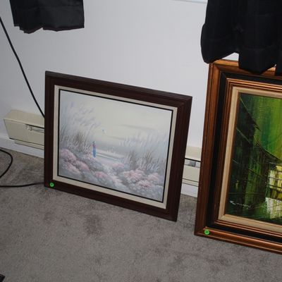 Sale Photo Thumbnail #72: Two framed vintage oil paintings resting on the floor, including a muted coastal landscape with tall grass and a mid-century modern green architectural cityscape.