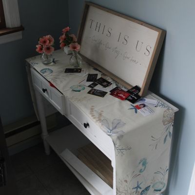 Sale Photo Thumbnail #30: White wooden entryway console table featuring two drawers and a lower shelf, topped with a coastal-themed table runner, small faux floral arrangements, and framed 'This is Us' wall art.