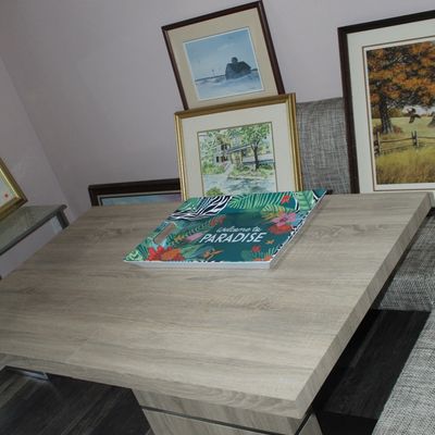 Sale Photo Thumbnail #13: Contemporary square light-wood dining table with pedestal base, displaying framed artwork and a tropical "Welcome to Paradise" tray.