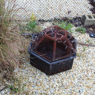 Sale Photo Thumbnail #66: Hexagonal outdoor metal fire pit with domed mesh spark screen topper, showing significant surface rust from exposure to weather.