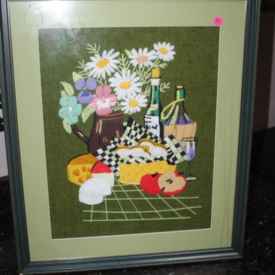Sale Photo Thumbnail #9: Framed vintage crewel embroidery piece depicting a still life picnic scene with flowers, wine, cheese, and fruit, set in a green mat and thin frame.