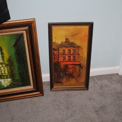 Sale Photo Thumbnail #74: Two framed vintage oil paintings on canvas, featuring a green monochrome cityscape on the left and a warm-toned European building scene signed Ricke on the right.
