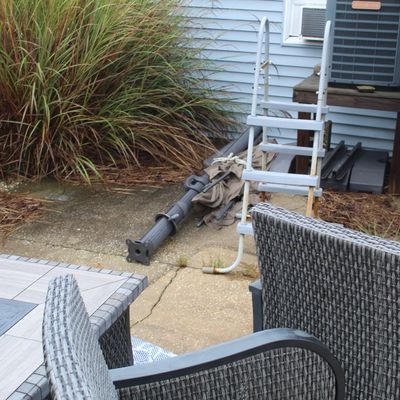 Sale Photo Thumbnail #55: Outdoor patio space featuring gray synthetic wicker armchairs, a matching tiled fire pit table, an A-frame pool ladder, and a collapsed outdoor umbrella.